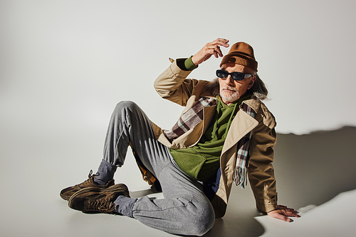 full length of expressive and trendy senior man in beanie hat, dark sunglasses, beige trench coat and sneakers sitting on grey background with shadow, hipster fashion, stylish lifestyle