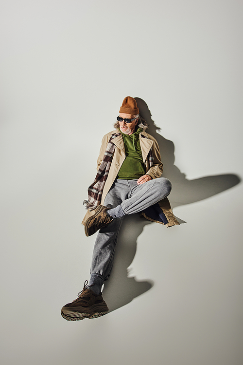 high angle view of senior male model in trendy hipster style attire and dark sunglasses laying and looking away on grey background with shadow, beanie hat, beige trench coat, fashion and age concept