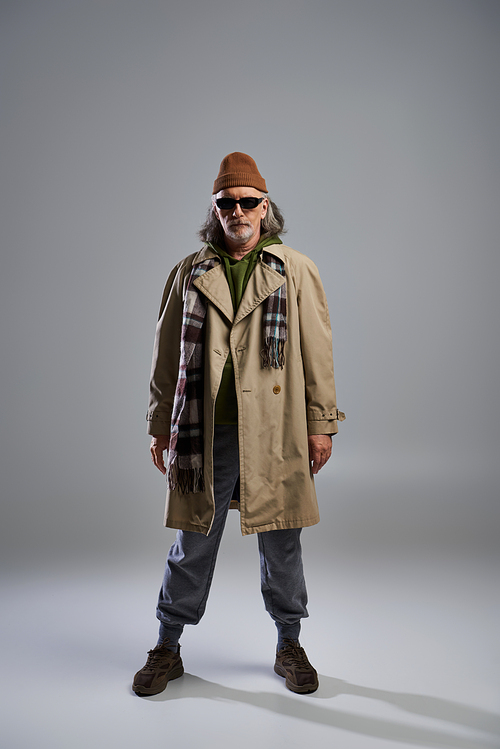 front view of trendy and bearded senior man in dark sunglasses, beanie hat, beige trench coat and sneakers standing on grey background, hipster fashion, trendy aging concept, full length