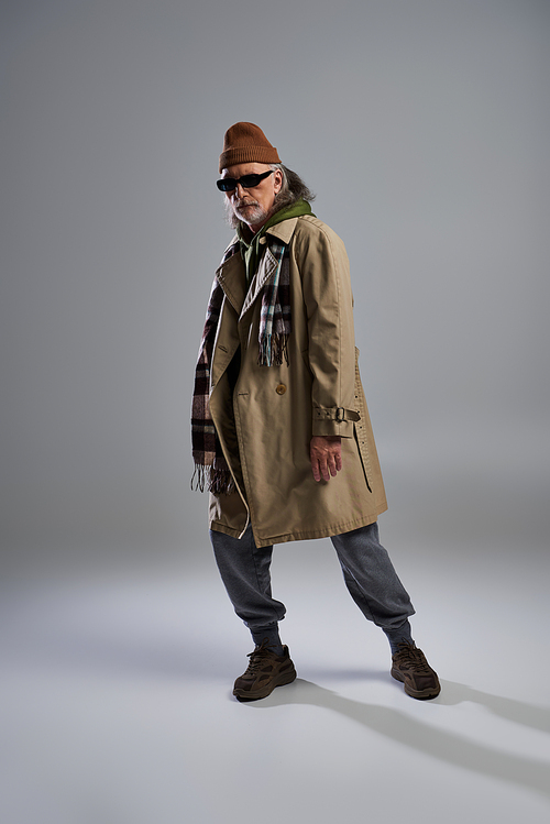 full length of senior hipster man in dark sunglasses and fashionable casual clothes standing and looking at camera on grey background, expressive personality, stylish aging concept