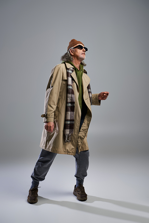 full length of aged and trendy man in beanie hat, dark sunglasses, beige trench coat and plaid scarf looking away while standing on grey background, stylish pose, fashion shoot