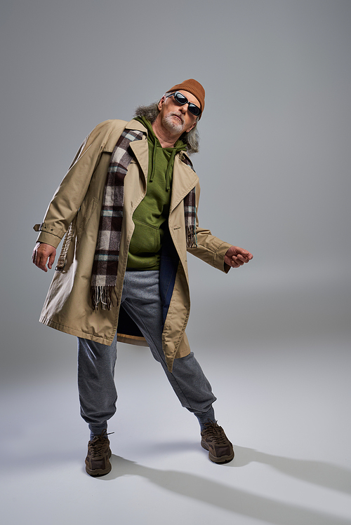 full length of aged senior model in hipster style outfit posing on grey background, beanie hat, dark sunglasses, beige trench coat, sneakers, fashionable lifestyle and positive aging concept