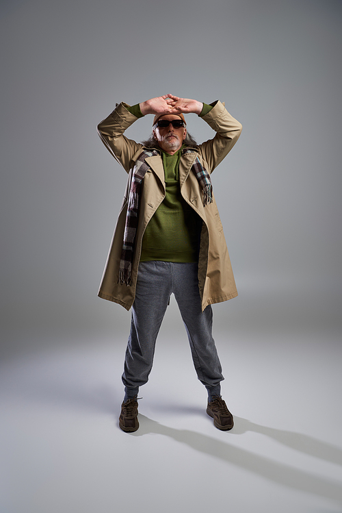 full length of cool and senior hipster man in dark sunglasses, beanie hat, beige trench coat and sneakers posing with hands above head on grey background, fashionable aging concept