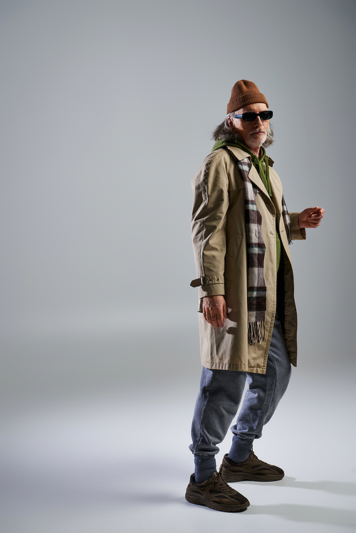 full length of senior and trendy man in dark sunglasses looking at camera on grey background, beanie hat, beige trench coat, plaid scarf, sneakers, fashion and age concept
