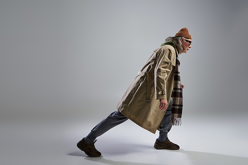 side view of senior and trendy model in hipster style outfit and dark sunglasses stepping on grey background with copy space, beanie hat, beige trench coat, sneakers, full length view, fashion shoot