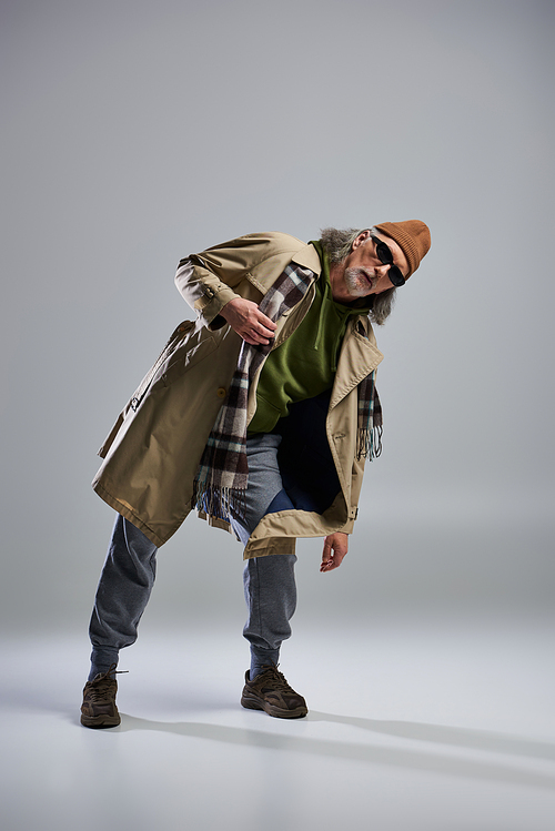 full length of expressive senior man in dark sunglasses, beanie hat, beige trench coat and plaid scarf looking at camera on grey background, hipster fashion, stylish pose, trendy aging concept