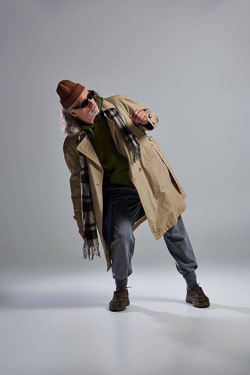 full length of elderly and bearded man in dark sunglasses, beanie hat, beige trench coat and plaid scarf standing in stylish pose and looking away on grey background