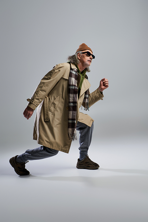 full length of grey haired man in dark sunglasses and hipster style outfit posing and looking away on grey background, beanie hat, beige trench coat, sneakers, expressive personality
