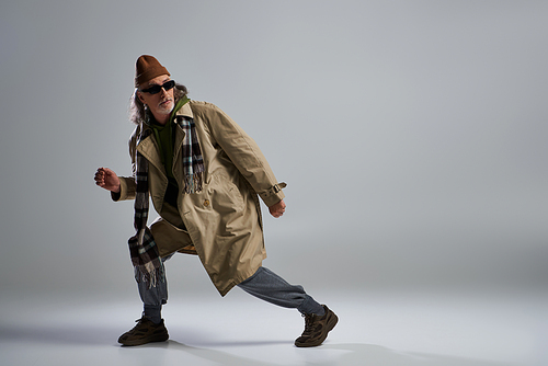 full length of fashionable senior man looking away on grey background, stylish pose, hipster, dark sunglasses, beanie hat, beige trench coat, stylish aging of expressive personality