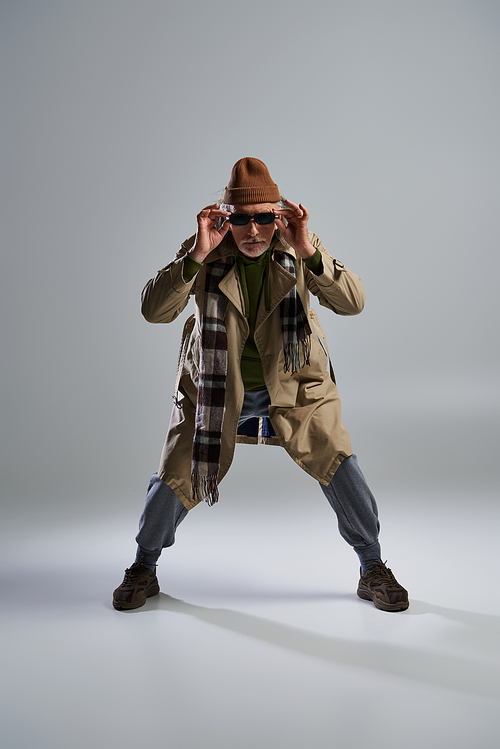 full length of fashionable senior model in stylish pose, aged hipster man in beanie hat and beige trench coat adjusting dark sunglasses and looking at camera on grey background