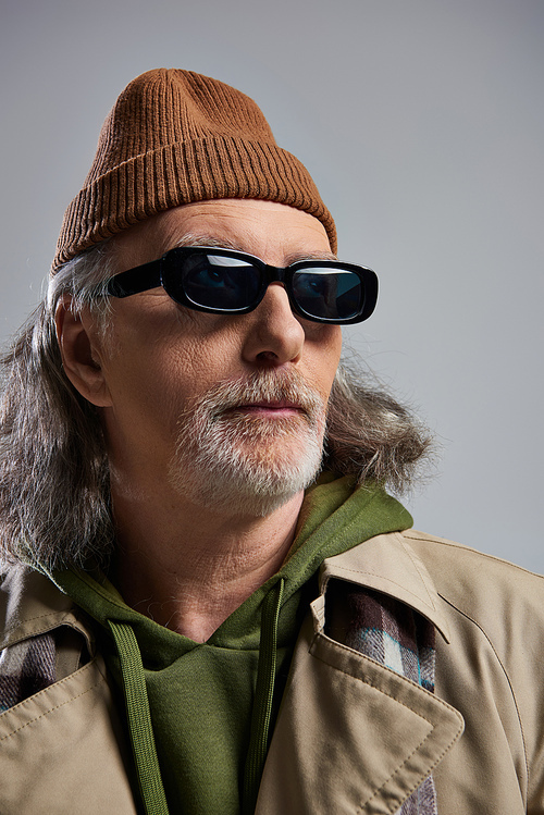 portrait of confident grey haired and bearded senior man in beanie hat, dark sunglasses and beige trench coat looking away on grey background, hipster style, fashion and age concept