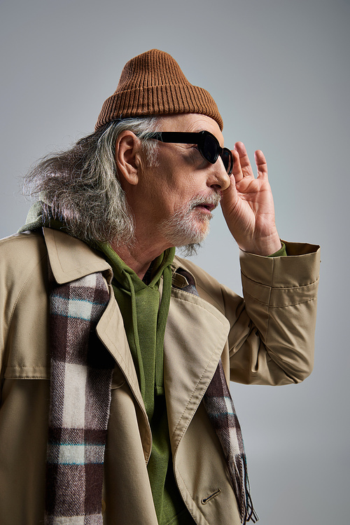 portrait of amazed elderly man in beanie hat, beige trench coat and plaid scarf adjusting dark sunglasses and looking away on grey background, hipster fashion, stylish and positive aging concept