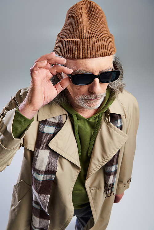 fashionable and bearded man adjusting dark sunglasses while standing on grey background, hipster fashion, beanie hat, beige trench coat, plaid scarf, aging with style concept, fashion shoot