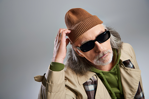 portrait of thoughtful elderly man in dark sunglasses and beige trench coat touching beanie hat and looking at camera on grey background, hipster fashion, stylish and positive aging concept