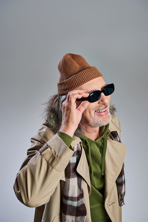 portrait of senior and cheerful hipster style bearded man in beanie hat and beige trench coat adjusting dark sunglasses, laughing and looking away on grey background