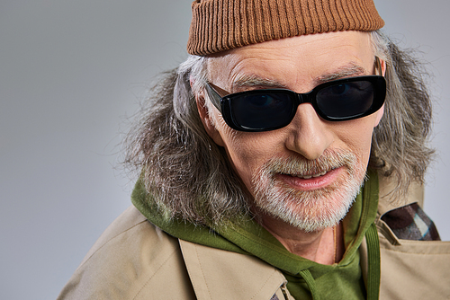 portrait of smiling senior man with grey hair and groomed beard, in dark sunglasses, beanie hat and trench coat looking at camera on grey background, hipster fashion, happy and trendy aging concept