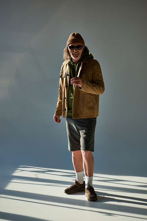 full length of cheerful senior man in beanie hat, dark sunglasses, jacket and shorts smiling at camera on grey background with lighting, hipster style, happy and fashionable aging concept