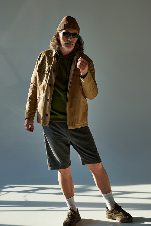 full length of aged bearded man in beanie hat, dark sunglasses, jacket and shorts looking at camera while standing on grey background with lighting, hipster fashion, trendy aging concept
