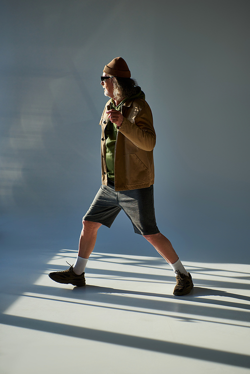 full length of senior man in hipster style outfit and dark sunglasses walking on grey background with lighting, beanie hat, jacket, shorts, trendy casual clothes, stylish aging concept