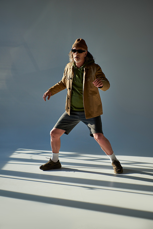full length of expressive senior man in trendy hipster clothes and dark sunglasses standing and posing on grey background with lighting, stylish pose, positive and fashionable lifestyle concept