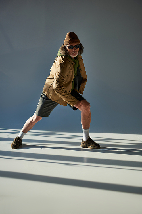 expressive and trendy personality, full length of senior man in dark sunglasses, beanie hat, jacket and shorts looking at camera while posing on grey background with lighting