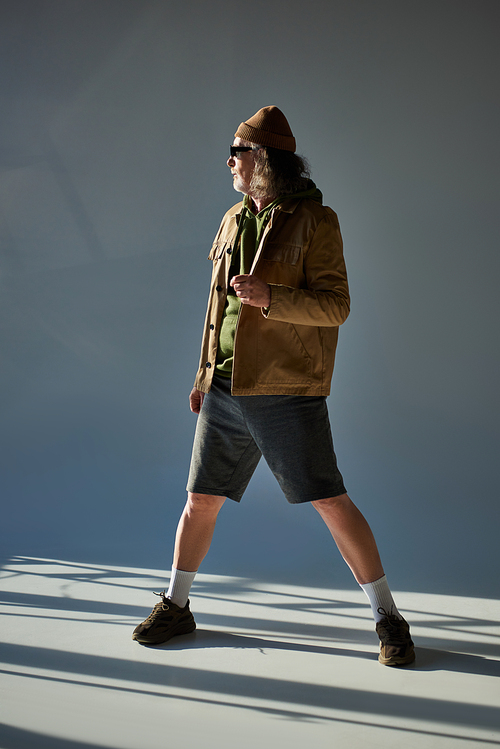 full length of elderly and grey haired man in beanie hat, dark sunglasses, jacket and shorts standing and looking away on grey background with lighting, hipster style aging concept