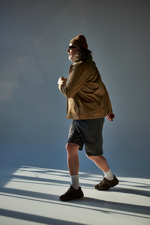 side view of bearded and hipster style man in fashionable casual clothes walking on grey background with lighting, beanie hat, dark sunglasses, jacket and shorts, fashion and age concept