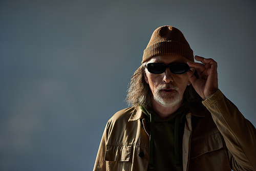 fashionable and hipster style senior man in beanie hat and brown jacket adjusting dark sunglasses and looking at camera on grey background, aging population lifestyle concept