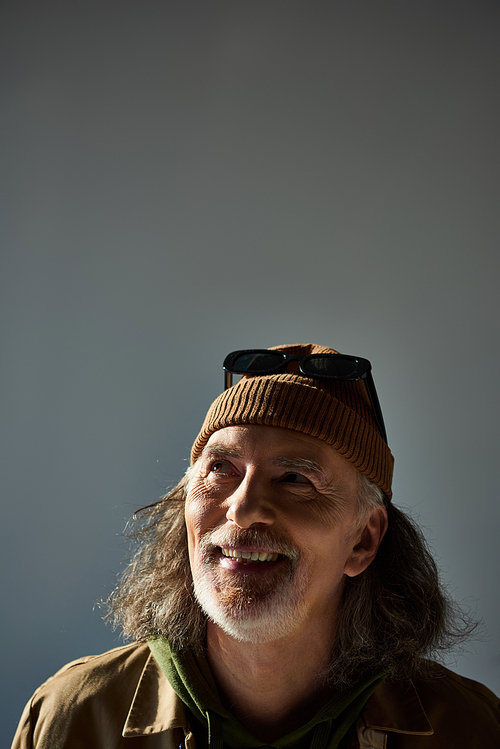 portrait of cheerful and carefree senior man with grey hair and beard, wearing dark sunglasses on beanie hat, looking up on grey background, with copy space, happy and fashionable aging concept