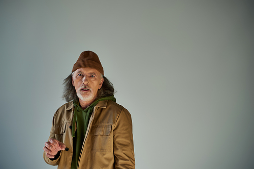 frustrated and worried elderly man with grey hair and beard, in beanie hat and brown jacket looking at camera on grey background, hipster style, aging population lifestyle concept