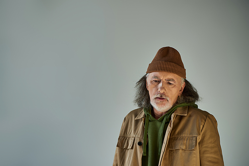 upset grey haired and bearded senior man in beanie hat and brown jacket standing on grey background, hipster style, expressive personality, aging population lifestyle concept