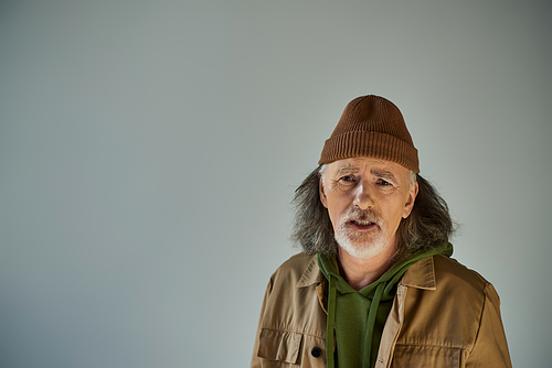 upset senior man with offended face expression looking at camera on grey background, grey hair, beard, beanie hat, brown jacket, hipster fashion, aging population lifestyle concept