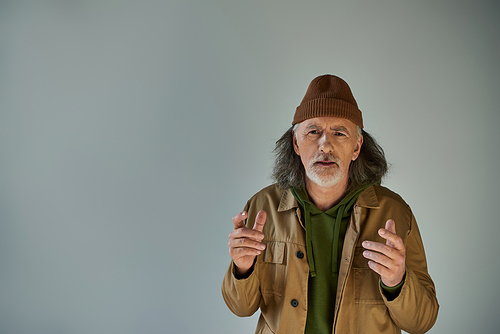 upset and worried senior bearded man in beanie hat and brown jacket gesturing and looking at camera on grey background, hipster clothes, aging population lifestyle concept