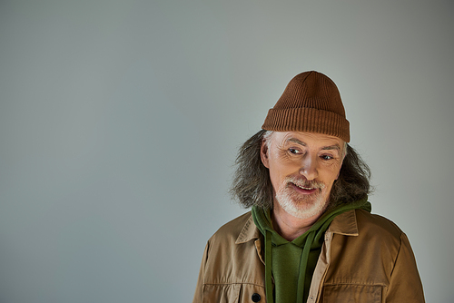 grey haired and bearded senior man, thoughtful and smiling, in beanie hat and brown jacket on grey background, hipster fashion, happy and fashionable aging concept