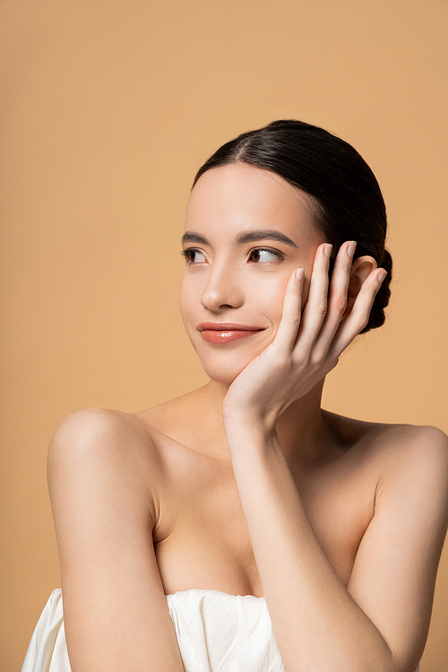 Pretty young asian woman with naked shoulders touching cheek while standing isolated on beige