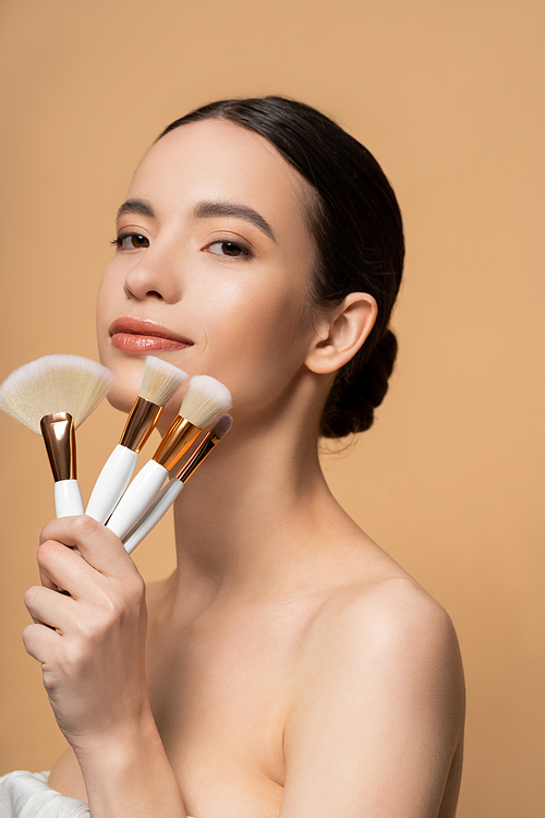Asian woman with naked shoulders holding makeup brushes and looking at camera isolated on beige