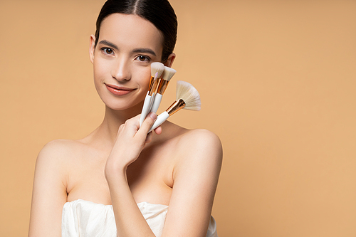 Pretty asian woman with naked shoulders in top holding makeup brushes and smiling isolated on beige