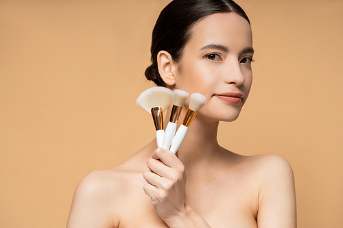 Brunette asian woman with naked shoulders holding makeup brushes isolated on beige