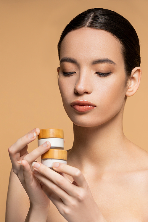 Young asian woman with naked shoulders and perfect skin holding cosmetic cream isolated on beige