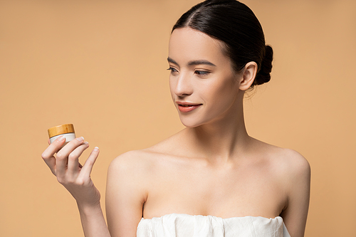 Positive young asian woman in top holding cosmetic cream while standing isolated on beige