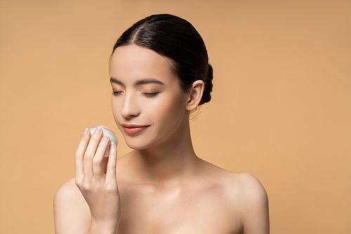 Young asian woman with naked shoulders smelling cosmetic cream while standing isolated on beige