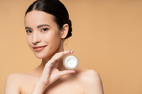 Happy asian woman with naked shoulders and natural makeup holding cosmetic cream isolated on beige