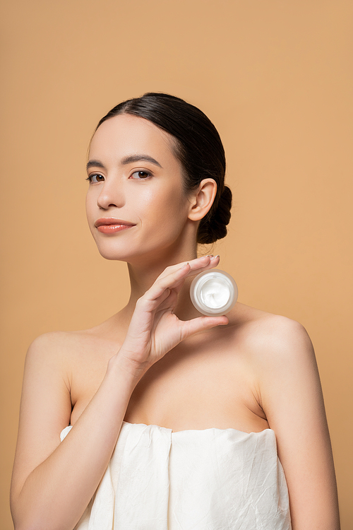 Young asian woman in top with naked shoulders holding cosmetic cream isolated on beige