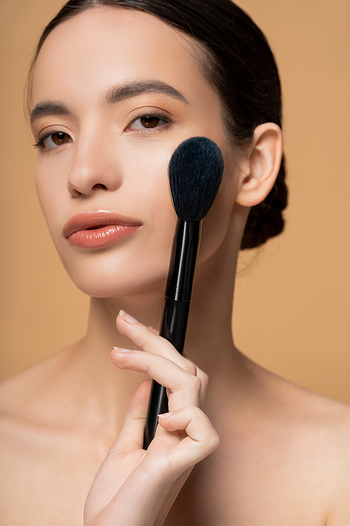 Young asian woman with natural makeup and naked shoulders holding blush brush isolated on beige