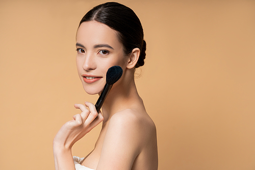Smiling young asian woman with naked shoulders holding blush brush near face isolated on beige
