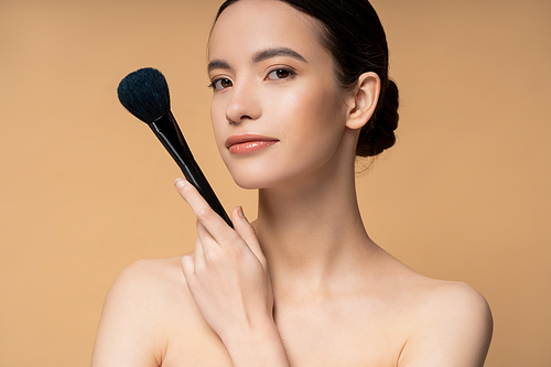 Confident young asian woman with naked shoulders holding makeup brush and standing isolated on beige