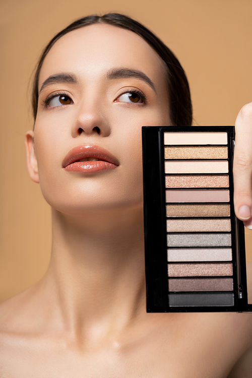 Young asian woman with natural makeup holding eyeshadow palette and looking away isolated on beige