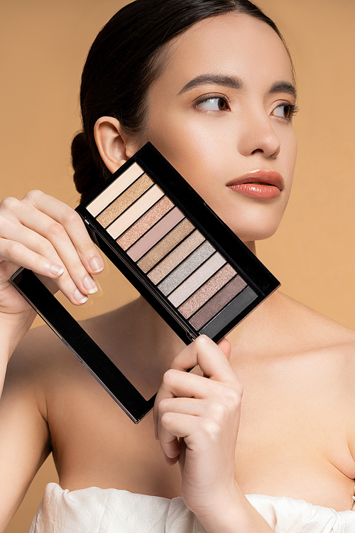 Asian model with perfect skin holding eyeshadow makeup palette while posing isolated on beige