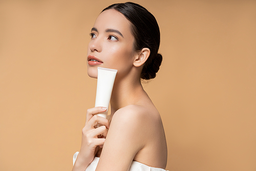 Young asian woman in top holding cosmetic lotion and looking away isolated on beige