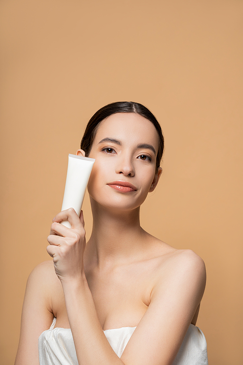 Portrait of young asian model in top holding cosmetic balm and looking at camera isolated on beige